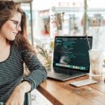 Woman sits at a wooden table in a cafe, looking at a laptop displaying code, with a drink and plants beside her.