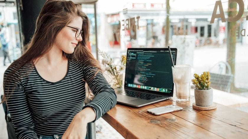 Woman sits at a wooden table in a cafe, looking at a laptop displaying code, with a drink and plants beside her.
