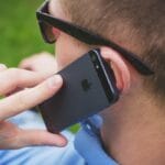 A person wearing sunglasses holds an iPhone to their ear while talking on the phone outdoors.