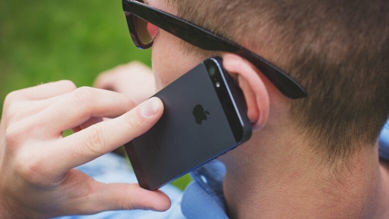 A person wearing sunglasses holds an iPhone to their ear while talking on the phone outdoors.