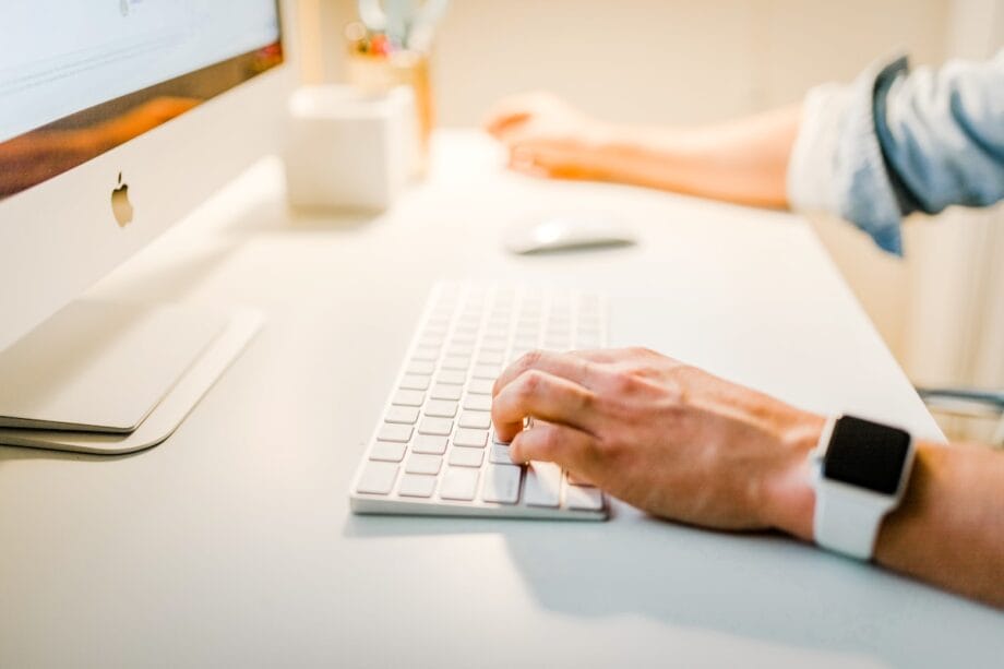 Person typing on a white keyboard at a desk with a computer monitor and wearing a smartwatch.