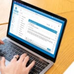 Person typing an email on a laptop with a blue and white interface, seated at a light wooden table.