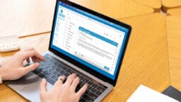 Person typing an email on a laptop with a blue and white interface, seated at a light wooden table.