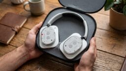 Hands holding an open case containing a pair of over-ear headphones with a transparent back cover on a wooden table.