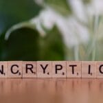 Scrabble tiles on a wooden surface spelling ENCRYPTION, with a blurred green and white background.