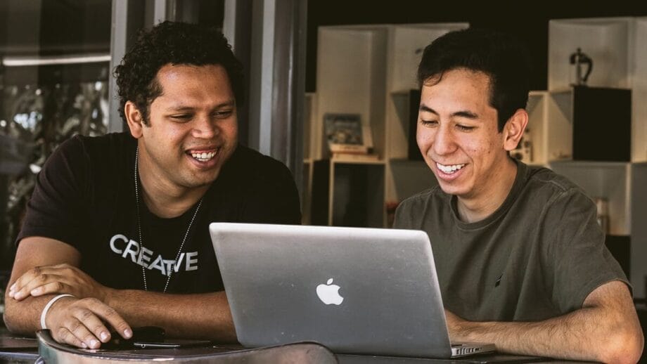 Two young men sit together at a table, smiling while looking at an open laptop in front of them.