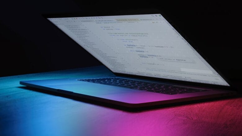 A partially open laptop displaying code on the screen, illuminated by blue and pink lighting on a dark surface.