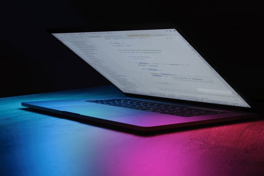 A partially open laptop displaying code on the screen, illuminated by blue and pink lighting on a dark surface.