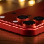 Close-up of a red smartphone with dual rear cameras and a flash, placed on a wooden surface with blurred lights in the background.