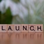 Wooden letter tiles on a wooden surface spell out the word LAUNCH with a blurred green and white background.