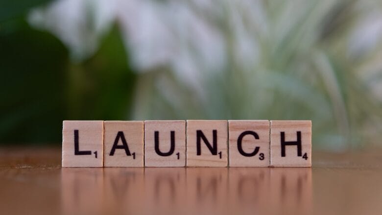 Wooden letter tiles on a wooden surface spell out the word LAUNCH with a blurred green and white background.