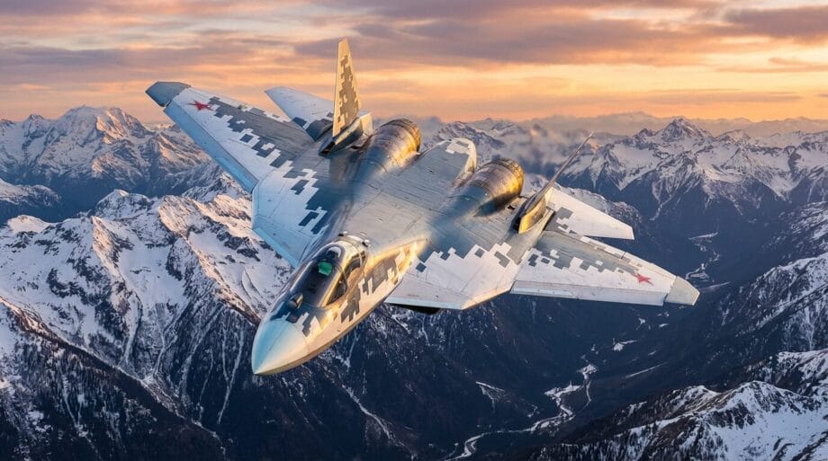 A military jet with a digital camouflage pattern flies over snow-covered mountains during sunset.