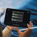 Person holding a stylus and using a tablet with a dark-themed app interface displaying a playlist or series of media options.