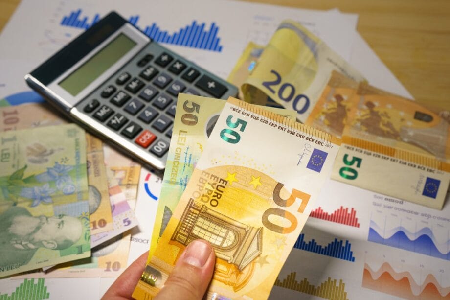 A hand holds euro banknotes over financial charts, with more euro bills and a calculator nearby on a desk.