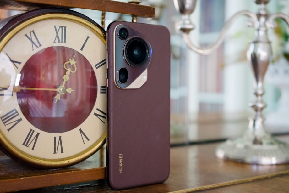 A Huawei smartphone leans against a vintage clock displaying 2:31, with a candlestick in the background on a wooden surface.
