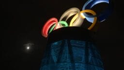 The Olympic rings are illuminated atop a blue structure at night, with the moon visible in the dark sky.