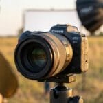 A Canon EOS R5 camera on a tripod is set up outdoors, with lighting equipment and two people visible in the background.