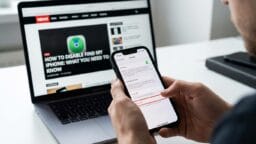 Person holding an iPhone with settings open, sitting in front of a laptop displaying a tech article about disabling Find My iPhone.
