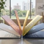 Six Apple MacBook Air laptops in different colors are arranged in a semicircle on a table in a bright, modern office.