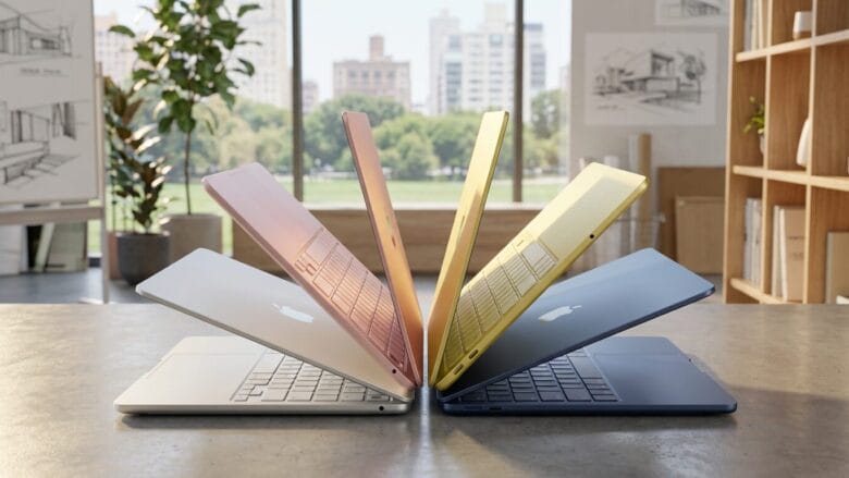 Six Apple MacBook Air laptops in different colors are arranged in a semicircle on a table in a bright, modern office.