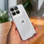 A hand holding a silver Motorola smartphone with four rear cameras, positioned indoors near a wooden table.