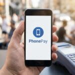 A person holds a smartphone displaying the PhonePay app near a payment terminal on an outdoor table.