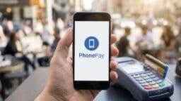 A person holds a smartphone displaying the PhonePay app near a payment terminal on an outdoor table.