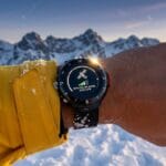 A person wearing a yellow jacket holds out a smartwatch displaying a satellite connection screen, with snowy mountains in the background.