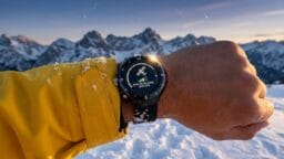 A person wearing a yellow jacket holds out a smartwatch displaying a satellite connection screen, with snowy mountains in the background.