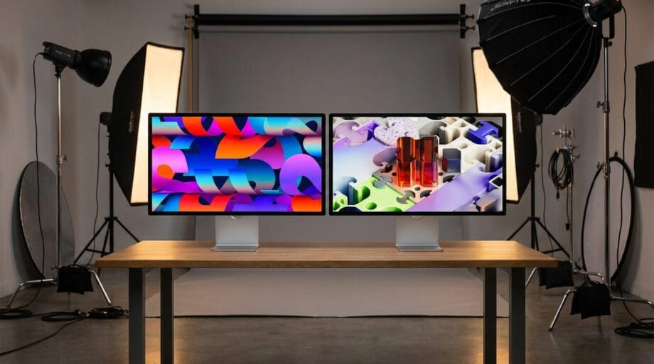 Two computer monitors on a wooden desk in a photography studio with professional lighting equipment in the background.