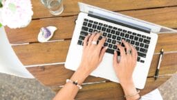 Person typing on a laptop at a wooden table with a pen, glass of water, and decorative items nearby.