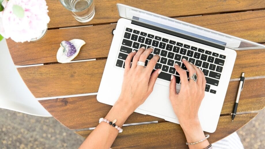 Person typing on a laptop at a wooden table with a pen, glass of water, and decorative items nearby.