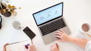 Two people work together at a desk with a laptop, notebook, pen, phone, and two coffee mugs on a white surface.