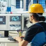 Engineer wearing a yellow hard hat works at a desk with two monitors displaying technical and structural diagrams in an office setting.