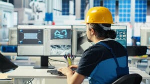 Engineer wearing a yellow hard hat works at a desk with two monitors displaying technical and structural diagrams in an office setting.