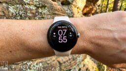 A person wearing a white smartwatch displaying the time 07:55, heart rate, and date, against a rocky outdoor background.