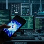 A person holds a smartphone with a lock icon in front of multiple computer screens displaying code in a server room.