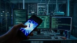 A person holds a smartphone with a lock icon in front of multiple computer screens displaying code in a server room.