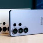 Two Samsung Galaxy S24 smartphones with triple rear cameras are displayed on a wooden surface, one silver and one cream colored.