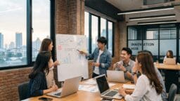 Six people in a modern office discuss charts and graphs on a whiteboard labeled STARTUP, with laptops on the table.