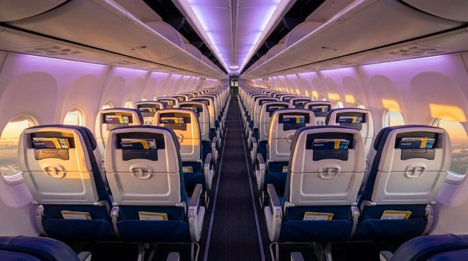 Empty airplane cabin with rows of seats and purple ambient lighting, viewed from the rear facing the front of the plane.