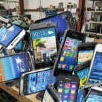 A pile of various old smartphones on a table in an electronics recycling facility with shelves and bins in the background.