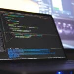 A laptop displaying lines of code on its screen, with another blurred monitor in the background.