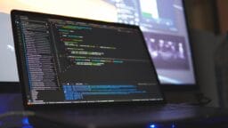 A laptop displaying lines of code on its screen, with another blurred monitor in the background.