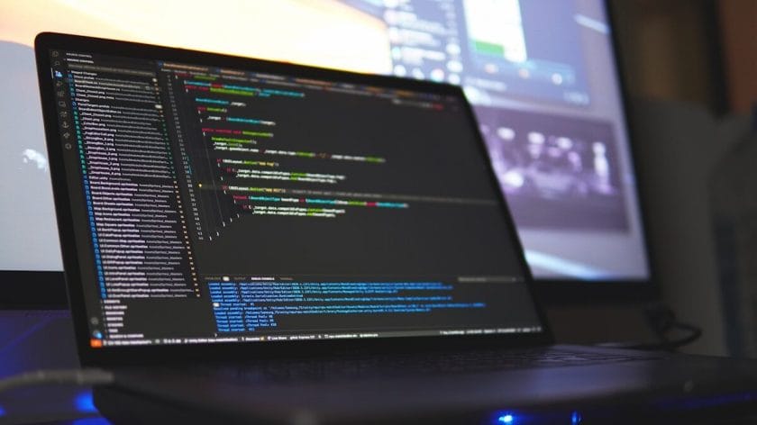 A laptop displaying lines of code on its screen, with another blurred monitor in the background.