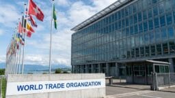 Flags from various countries stand outside the World Trade Organization building on a partly cloudy day.