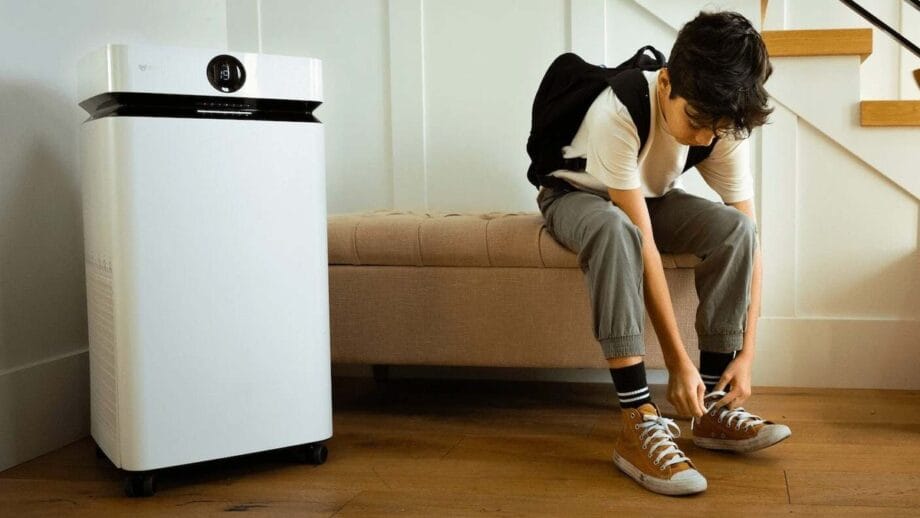 A boy with a backpack sits on a bench tying his shoes next to a white air purifier in a home setting.
