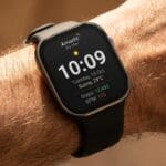 A smartwatch on a persons wrist displays the time, date, weather, step count, and heart rate on its home screen.