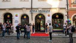 People gather outside the Apple Museum, with rainbow Apple logos and a red carpet at the entrance, as photographers take pictures.