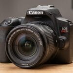 Canon EOS Rebel SL3 DSLR camera with an 18-55mm lens resting on a wooden surface, background out of focus.